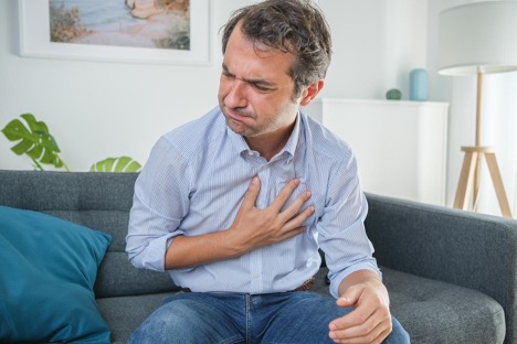 The man sitting on the sofa, holding his chest, is suffering from gastroesophageal reflux after eating
